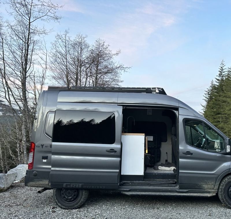 Van parked on gravel with sliding door open by the water and tall evergreens