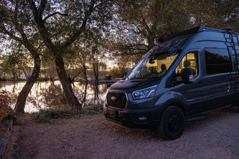 The van parked under oak trees beside a still lake at dusk